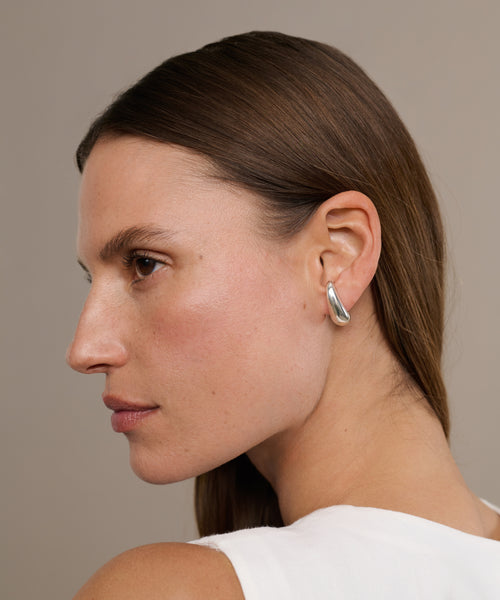 A woman with straight brown hair, wearing a sleeveless white top and Noodle Earrings, is shown in profile against a neutral background.