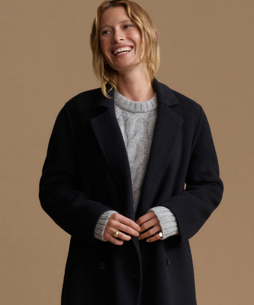 A woman smiling and wearing a black wool coat over a light gray cable-knit sweater, posed against a solid warm beige studio background.