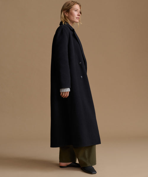 A woman stands in profile wearing a long black coat over olive green wide-leg pants and black flats, photographed against a plain warm beige studio background.