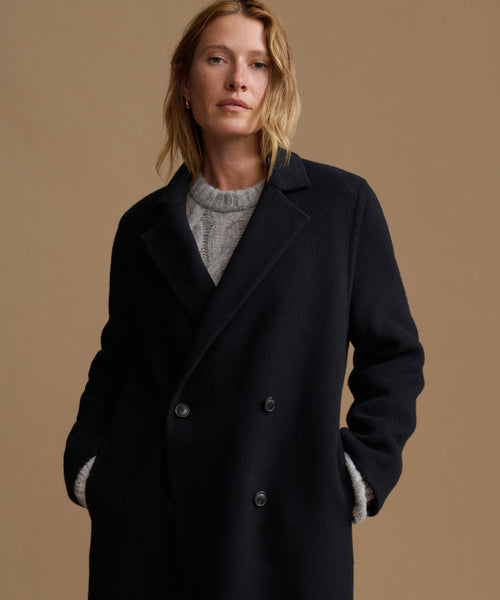 A woman wearing a black double-breasted coat over a light gray knit sweater, standing against a solid warm beige background.
