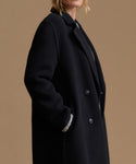 A close-up side view of a model wearing a black wool-blend coat with a double-breasted button closure and a ribbed knit collar detail, set against a solid warm tan background.