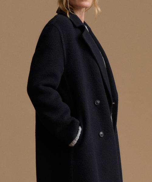 A close-up side view of a model wearing a black wool-blend coat with a double-breasted button closure and a ribbed knit collar detail, set against a solid warm tan background.