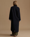 A model viewed from the back wearing a long, black wool-blend coat over olive green trousers, set against a solid warm beige studio background.