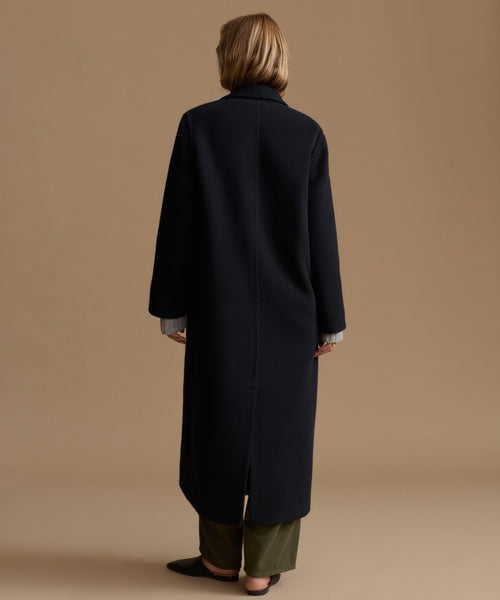 A model viewed from the back wearing a long, black wool-blend coat over olive green trousers, set against a solid warm beige studio background.