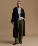 A woman stands against a plain beige studio background wearing a long dark black coat over a light gray cable-knit sweater, olive green wide-leg trousers, and black loafers.