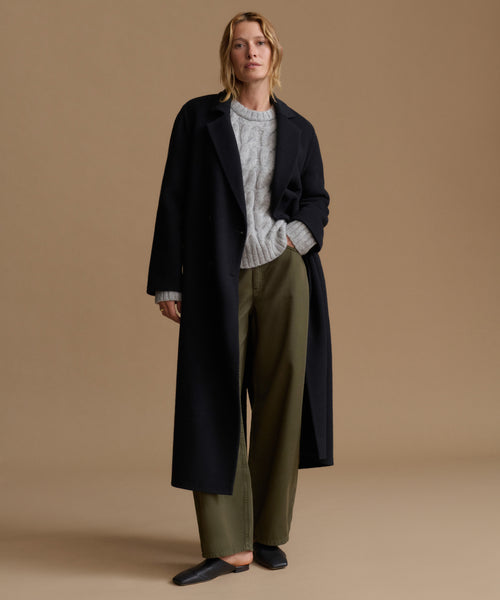 A woman stands against a plain beige studio background wearing a long dark black coat over a light gray cable-knit sweater, olive green wide-leg trousers, and black loafers.