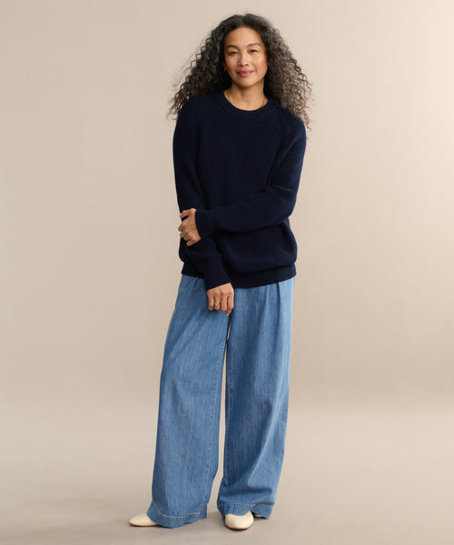 A woman with curly hair stands against a plain beige background, smiling gently with arms crossed while wearing the Oversized Cotton Fisherman sweater in navy, paired with light blue wide-leg jeans and white shoes.