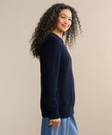 A woman with long curly hair smiles while standing sideways, wearing the Oversized Cotton Fisherman sweater in navy and light blue jeans against a neutral beige background.