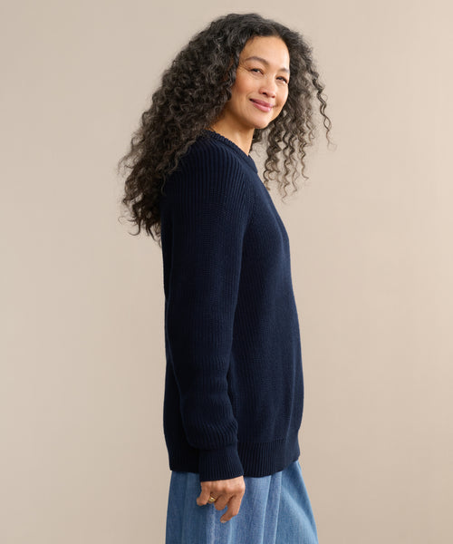 A woman with long curly hair smiles while standing sideways, wearing the Oversized Cotton Fisherman sweater in navy and light blue jeans against a neutral beige background.