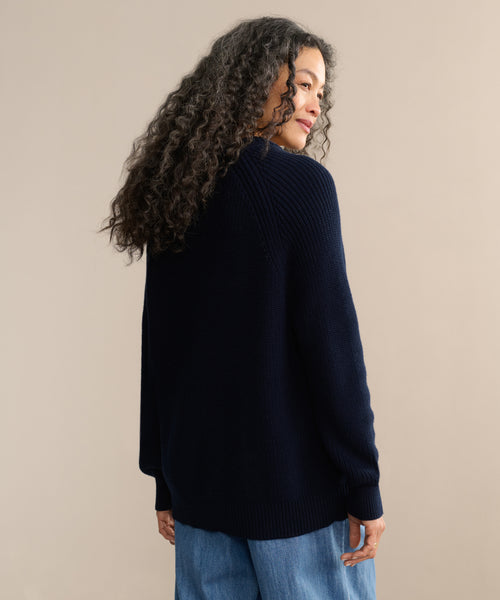 A person with long, curly hair wears the Oversized Cotton Fisherman sweater in navy with light blue jeans, standing against a plain beige background and looking over their shoulder.