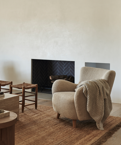 A comfortable Aspen Chair with a gray knitted throw sits on a brown textured rug by a dark fireplace. Nearby, two wooden stools and a stone coffee table complete the neutral-toned living room setting.
