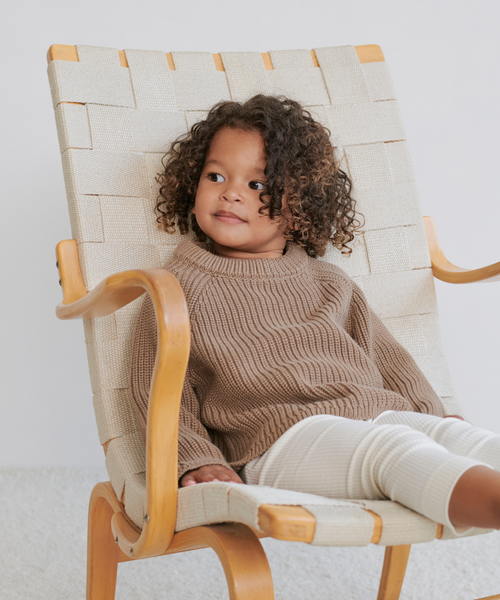 A young child with curly hair, dressed in the Kids Cotton Fisherman sweater and light pants, sits relaxed in a modern wooden chair with woven fabric, looking to the side and smiling gently.