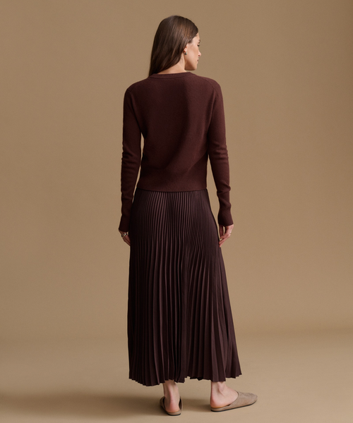 A woman is shown from behind wearing a dark brown long-sleeve knit top and a matching dark brown pleated midi skirt, paired with light brown flat shoes, against a solid tan studio background.