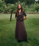 A woman stands on a grassy lawn wearing a dark brown short-sleeve collared top and matching pleated maxi skirt, with black ankle boots. Trees and shrubs are visible in the background.