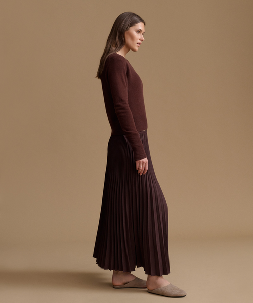 A woman in side profile wearing a dark brown long-sleeve knit top and a matching dark brown pleated maxi skirt, paired with taupe slip-on shoes, against a solid beige studio background.