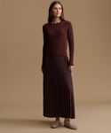 A woman stands against a plain warm beige studio background wearing a dark brown long-sleeve knit sweater and a matching dark brown pleated maxi skirt, with taupe slip-on shoes.
