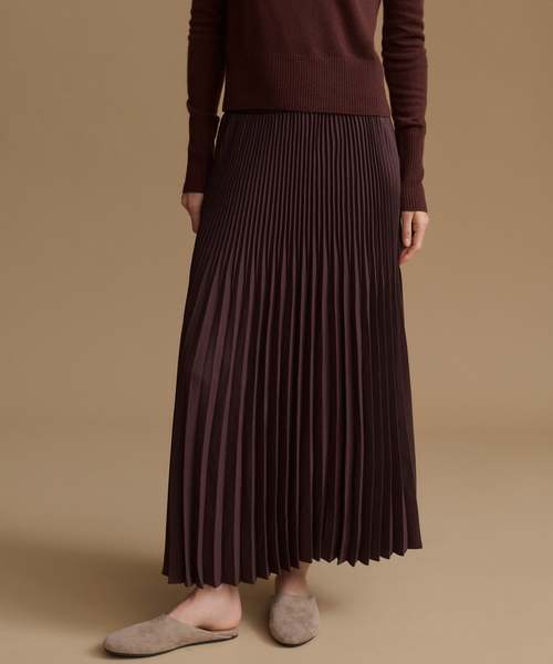 A person wearing a dark brown pleated maxi skirt with a ribbed knit top and taupe slip-on slippers, standing against a solid beige studio background.