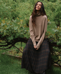 A woman stands outdoors in a lush green garden, wearing a beige turtleneck sweater and a dark gray pleated skirt with a brown plaid pattern.