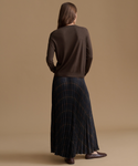 A woman is shown from behind wearing a dark brown long-sleeve knit sweater and a dark navy pleated maxi skirt with a subtle gold grid pattern, set against a solid warm beige studio background.