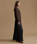 A woman stands in profile wearing a dark brown long-sleeve knit top and a dark navy pleated maxi skirt with a subtle diagonal stripe pattern, set against a solid warm beige studio background.