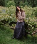 A woman posing outdoors in a lush green garden, wearing a beige knit sweater and a dark navy plaid maxi skirt.