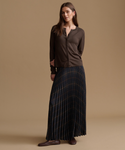 A woman modeling a dark brown long-sleeve button-front top and a dark pleated skirt with thin gold-toned stripes, standing against a plain beige studio background.