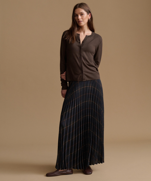 A woman modeling a dark brown long-sleeve button-front top and a dark pleated skirt with thin gold-toned stripes, standing against a plain beige studio background.