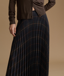 A close-up of a model wearing a dark navy pleated maxi skirt with a subtle brown and gold wavy stripe pattern, paired with a brown cardigan, against a solid beige background.