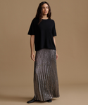 A woman stands against a plain warm tan studio background wearing a loose black short-sleeve top and a long pleated metallic taupe skirt with black shoes.