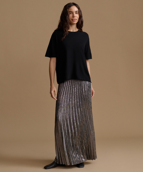 A woman stands against a plain warm tan studio background wearing a loose black short-sleeve top and a long pleated metallic taupe skirt with black shoes.