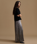 A woman stands in profile wearing a black short-sleeve top and a floor-length pleated metallic silver skirt against a plain warm beige studio background.