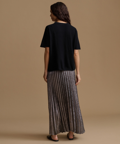 A woman is shown from behind wearing a black short-sleeve top and a pleated metallic gray maxi skirt, standing against a solid beige studio background.
