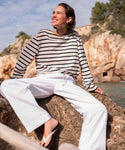 A woman in Paloma Pant summer pants, a striped long-sleeve shirt, and flip-flops smiles while sitting on a rock by the sea. Rocky cliffs, trees, and a partly cloudy sky create a scenic background.