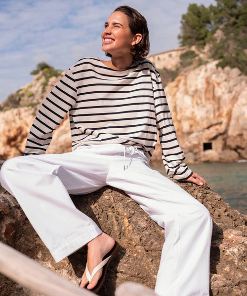 A woman in Paloma Pant summer pants, a striped long-sleeve shirt, and flip-flops smiles while sitting on a rock by the sea. Rocky cliffs, trees, and a partly cloudy sky create a scenic background.