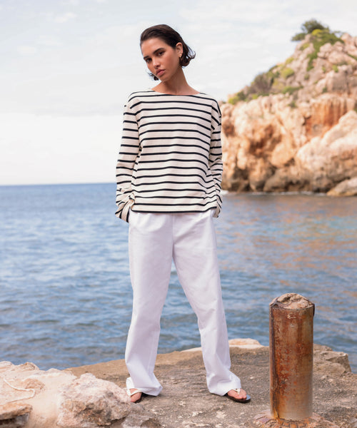 A person in a black-and-white striped long-sleeve shirt, Paloma Pant, and sandals stands on a rocky shore by the sea with cliffs and sparkling water in the background.