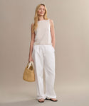A woman with long, wavy blonde hair wears a sleeveless beige top, Paloma Pant in loose white oxford fabric, and black sandals. She poses against a plain background, holding a woven straw bag.