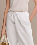A person wears a sleeveless white top and the Paloma Pant—loose heavyweight oxford summer pants—standing against a neutral background, with one hand in their pocket and holding a woven straw bag in the other.