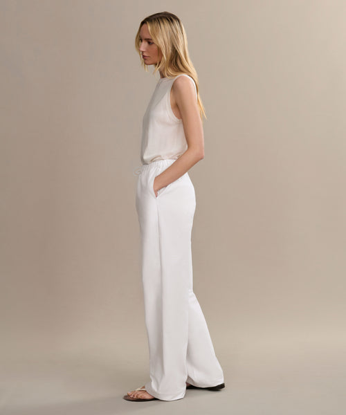 A woman in profile with long blonde hair, wearing a sleeveless white top and the Paloma Pant—our signature heavyweight oxford summer pant—stands with hands in pockets against a plain beige background.