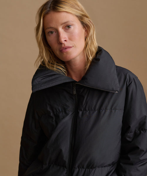 A close-up shot of a woman wearing a black puffer jacket with a high collar, set against a solid warm beige background.