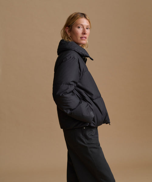A woman wearing a black puffer jacket and black pants, standing in profile against a solid beige studio background.
