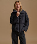 A woman modeling a black puffer jacket with a high collar and front zipper, paired with black pants, against a solid tan studio background.