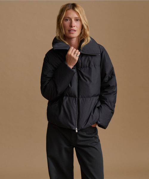 A woman modeling a black puffer jacket with a high collar and front zipper, paired with black pants, against a solid tan studio background.