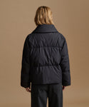 A model viewed from the back wearing a black puffer jacket and dark trousers, set against a solid warm tan studio background.