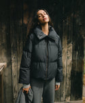 A woman wearing a black puffer jacket and gray sweatpants stands against a dark wooden plank wall, holding a gray coat in one hand.