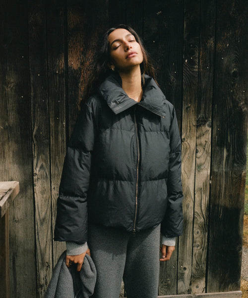 A woman wearing a black puffer jacket and gray sweatpants stands against a dark wooden plank wall, holding a gray coat in one hand.