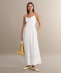 A woman stands against a plain beige background wearing the Peninsula Dress—a long white Italian washed poplin sundress with thin straps—paired with cream flat shoes and a woven straw handbag.