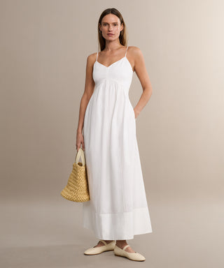 A woman stands against a plain beige background wearing the Peninsula Dress—a long white Italian washed poplin sundress with thin straps—paired with cream flat shoes and a woven straw handbag.
