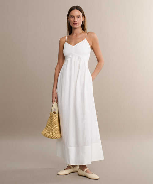 A woman stands against a plain beige background wearing the Peninsula Dress—a long white Italian washed poplin sundress with thin straps—paired with cream flat shoes and a woven straw handbag.
