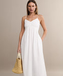 A woman in a white Peninsula Dress stands against a neutral background, holding a woven straw handbag. The sleeveless maxi boasts thin straps and is crafted from soft Italian washed poplin, offering effortless summer elegance.