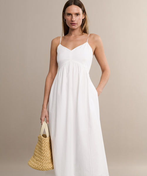 A woman in a white Peninsula Dress stands against a neutral background, holding a woven straw handbag. The sleeveless maxi boasts thin straps and is crafted from soft Italian washed poplin, offering effortless summer elegance.